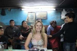 A white, blonde woman sits in a small-roomed teahouse. Behind her sit a row of adults - locals from Kathmandu. Next to her a young man dressed in black has his hand out and is talking to the adults.