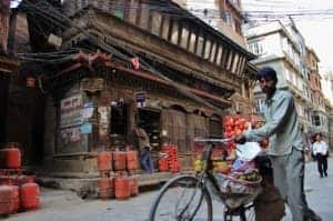 A man dressed in a grey suit, pushes his bicycle laden with shopping bags, past an old, dark brown wooden building in Kathmandu. Piles of orange canisters are stacked outside the building.