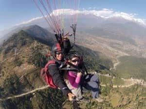 A woman sits in front of a male pilot in a paragliding harness, as they hover over a green valley backed by mountains in Pokhara, Nepal.