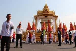 A parade of people walk alongside a tall, golden temple structure with a boat shaped base, carrying former Cambodian King Norodom Sihanouk in Phnom Penh at his state funeral