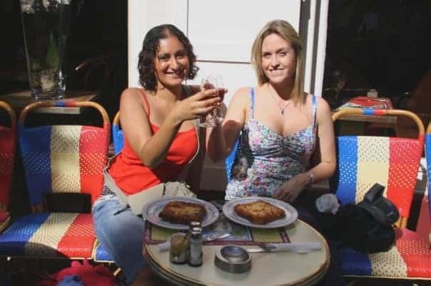Two women in a cafe in Paris, France making a toast with drinks. They are sitting on chairs made from fabric representing the French flag.