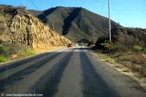 A wide, black and grey striped highway in sunny California. The view is from inside the car driving towards the golden and green high hills that line the road.