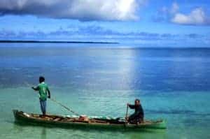 Two men, one standing and one sitting, row a long wooden boat on the turquoise waters backed by vast blue ocean.