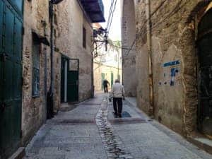 A man walks down a wide paved street lined either side with golden stone buildings with deep green doors, in the biblical city of Nazareth.