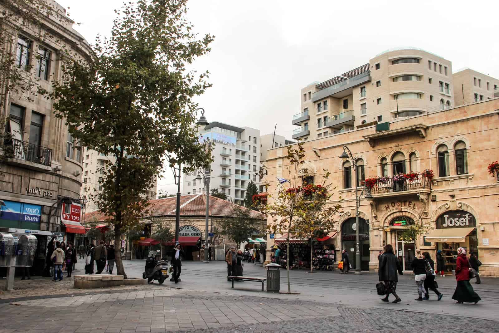 A small group of people walked past a caramel coloured low rise building in Jerusalem with the word "Hostel" over the arched doorway and a shop called Josep to the right. Behind it are two higher apartment buildings. 