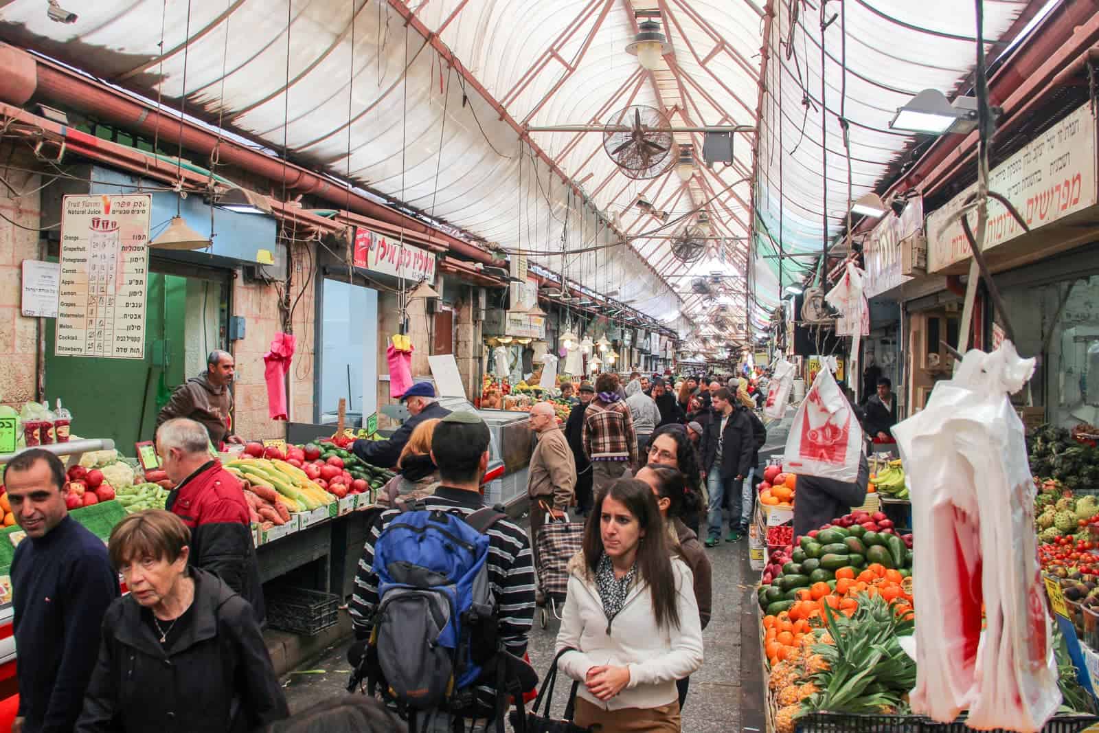 People walking. through the Jerusalem Mahaneh Yehuda Market, lined ether side with fresh fruits and vegetables and hanging plastic bags. The ceiling is covered, but lets in light. 