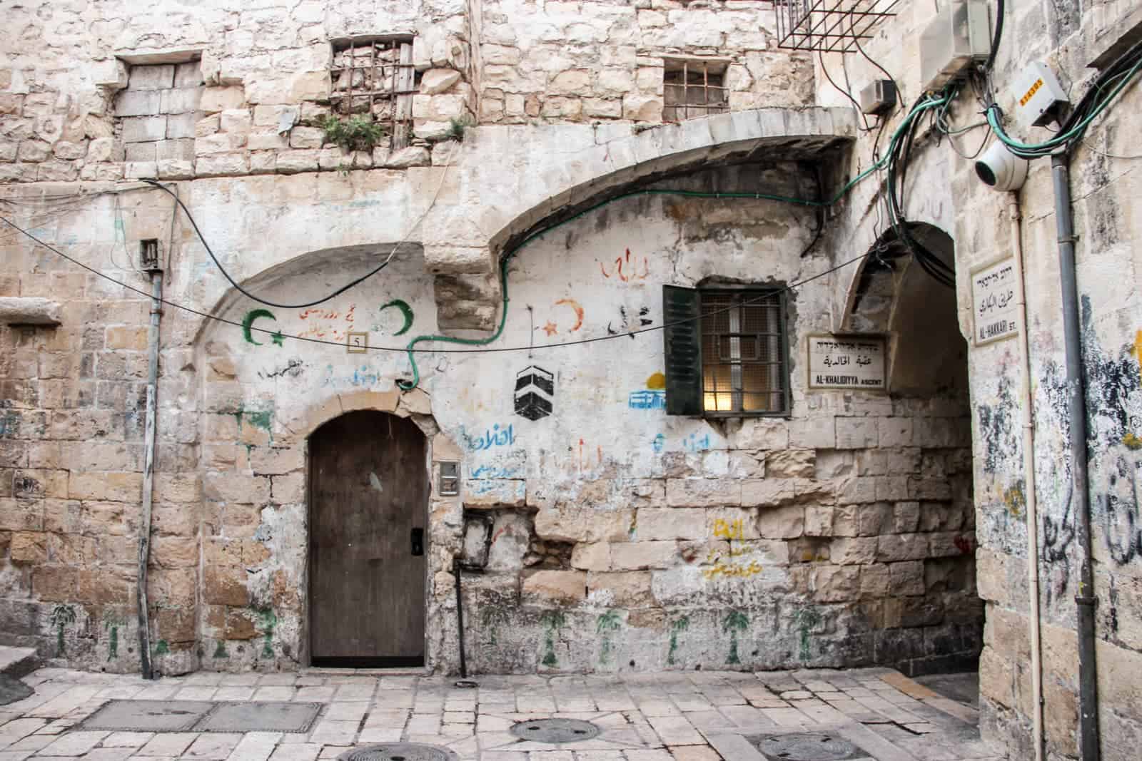 The worn old stone walls in Jerusalem's Old City covered with small scrolls of graffiti tags and religious symbols. A street name sign is next to the archway, on both sides of it. 