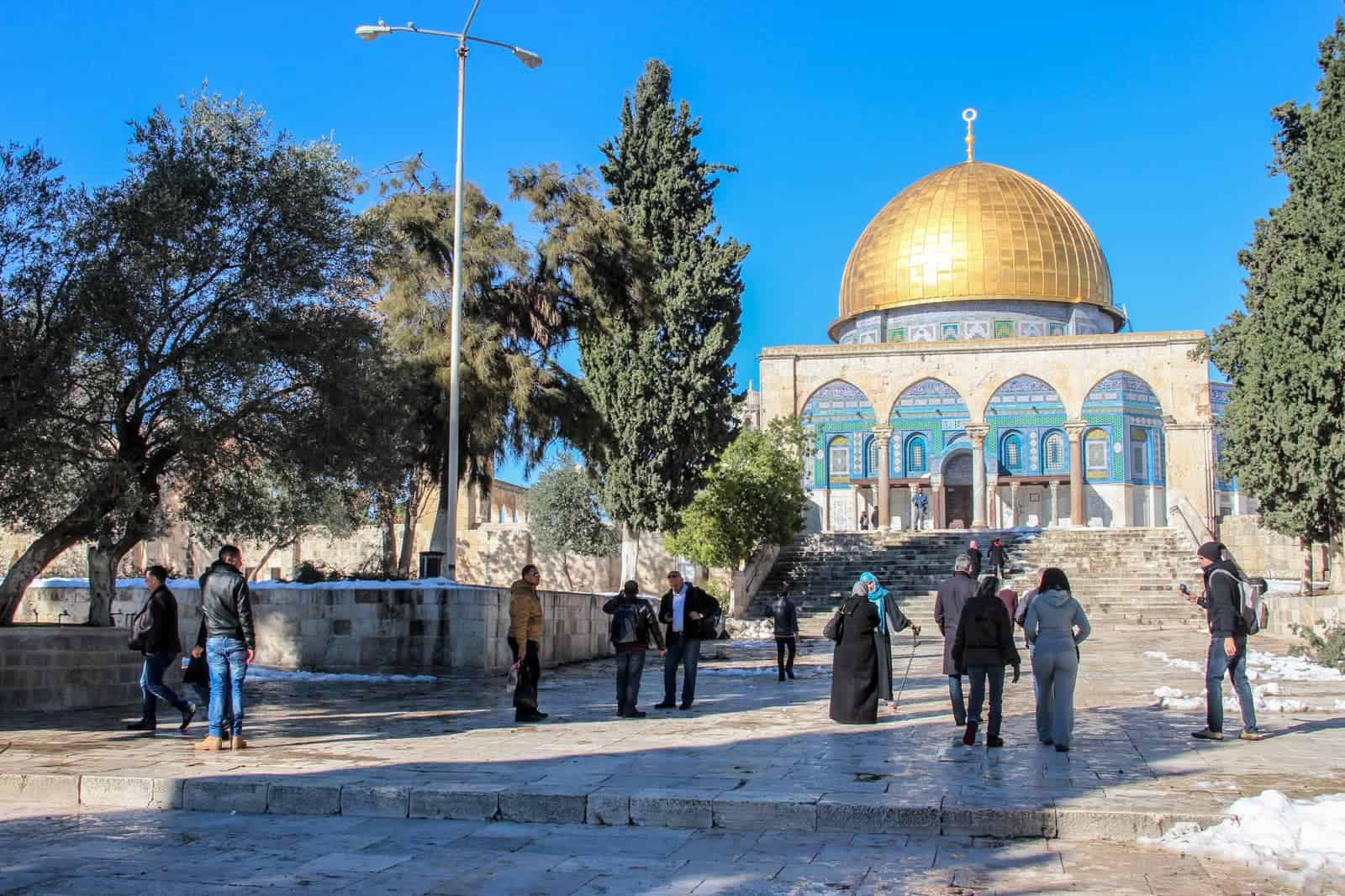 People walking towards a beige building with blue and green mosaics and a gold dome - Temple Mount in Jerusalem. Remnants of snow can be seen on the ground. 