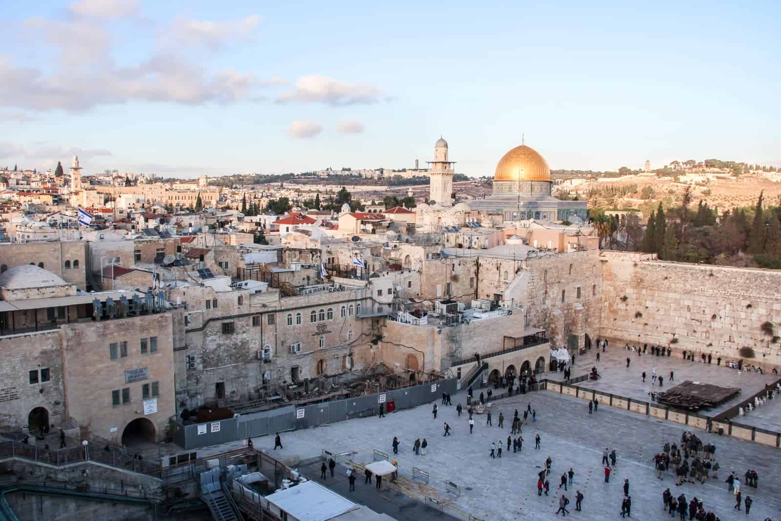 People gather in the The Western Wall Plaza in Jerusalem, surrounding by light orange-white stone walls and with a view to the golden dome of Temple Mount and a minaret.