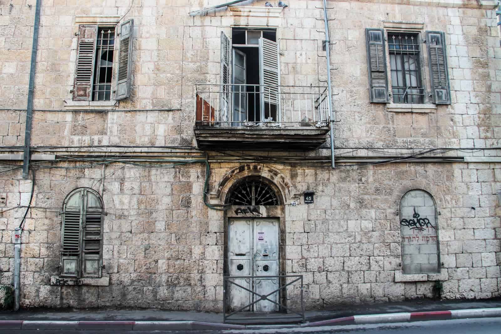 A worn Jerusalem stone building, with five windows (the bottom right one bricked up), and a grubby white metal door. 