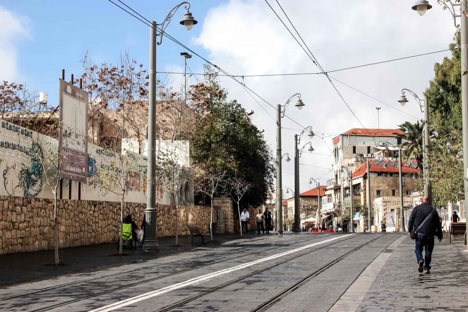 A man in dark clothing walks on the right side of the street, next to tram tracks in the road. To the left of him is a wall built with sandy coloured round stones and ahead, a cluster of buildings with orange roofs - the contemporary side of Jerusalem city