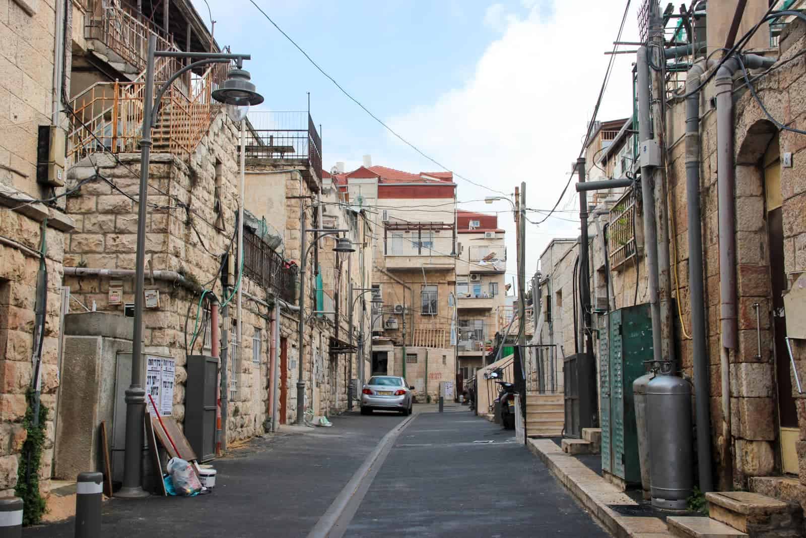 A local neighbourhood street in Jerusalem with low rise buildings built with neat honey coloured stones. A grey lamppost can be seen on the left and red rooftops in the background. 