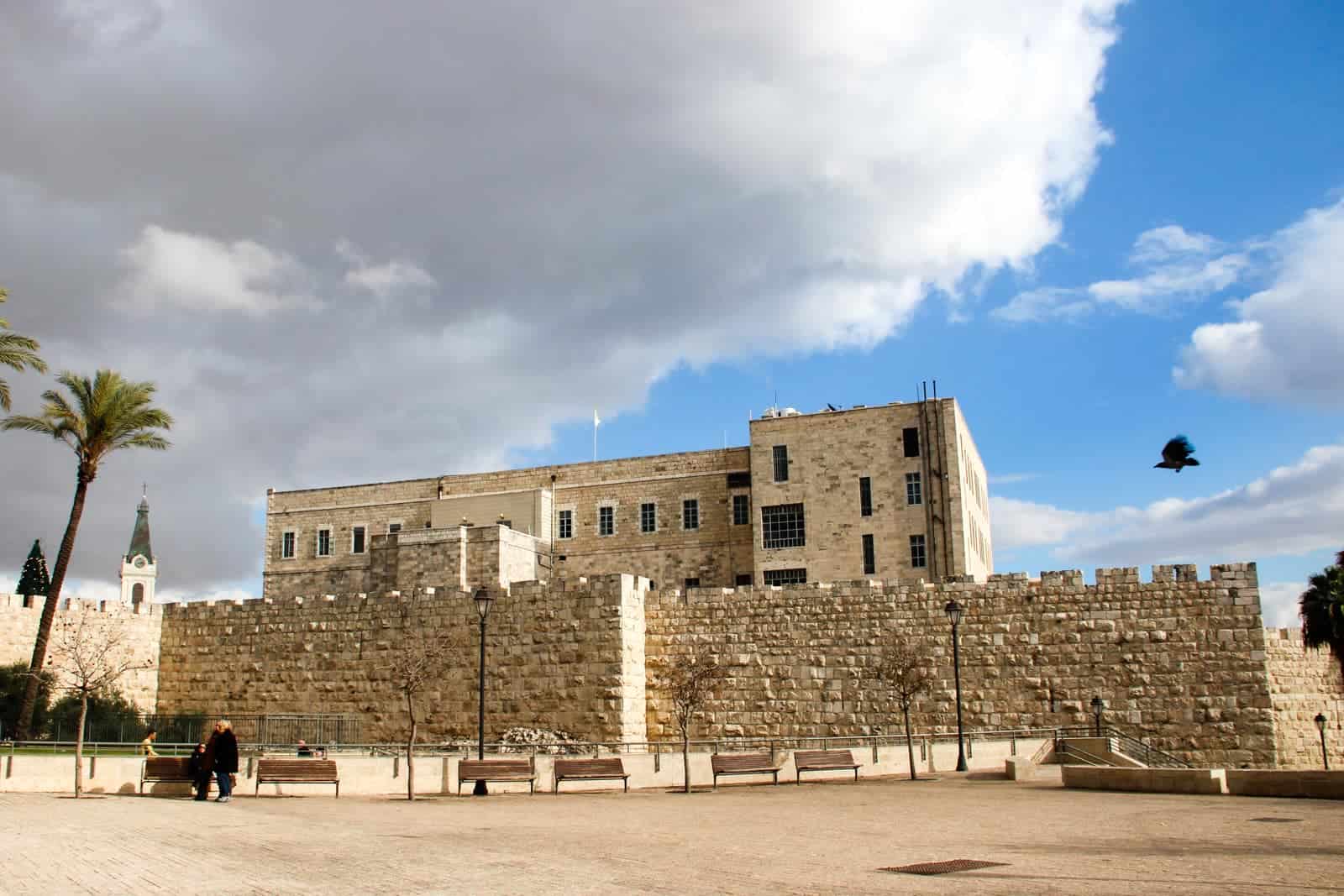 A man dressed in black sits on a bench in front of the glowing yellow of the Jerusalem Old City Walls that looks fortress like. A black bird flies towards the building. 