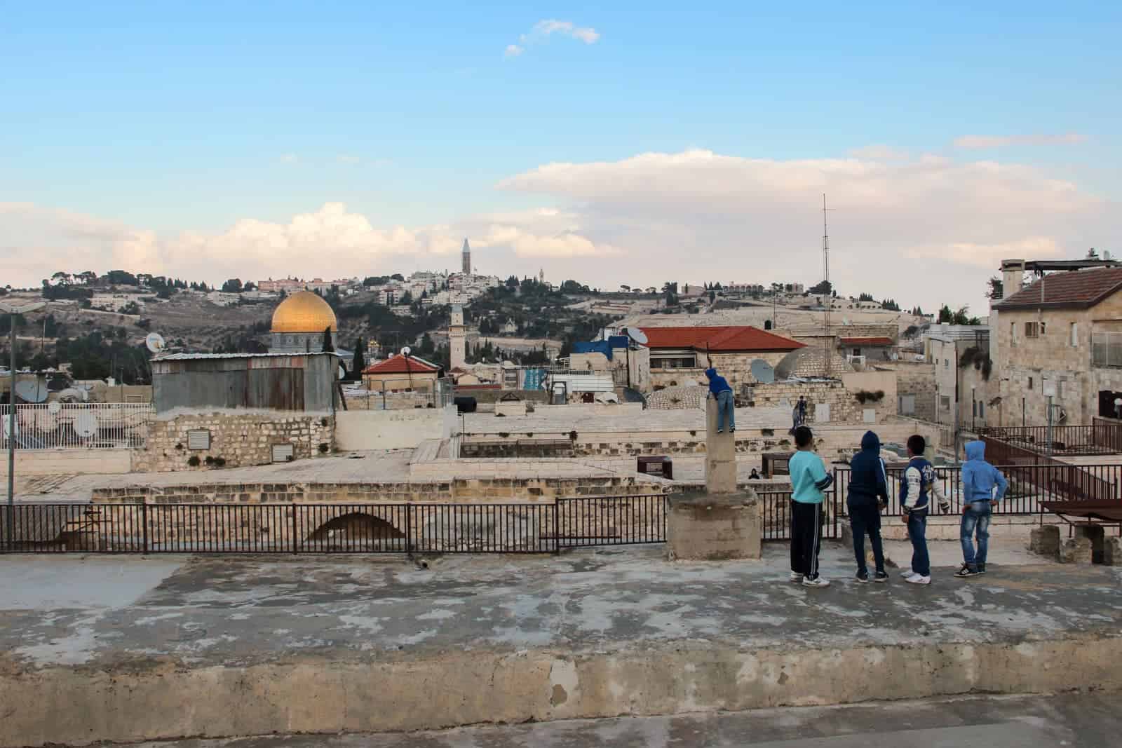 Four children standing on a gritty rooftop in Jerusalem overlooking a batch of beige stone buildings, the golden dome of a temple and out to the green olive groves on the far hills.