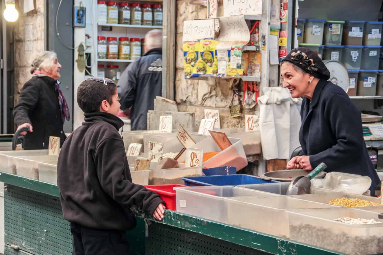 A female vendor at Jerusalem Mahaneh Yehuda Market in dark clothing and a head covering, smiles as she chats with a young boy, dressed in dark clothing and wearing a Jewish kippah. They are surrounded by boxes of spices and lentils. 