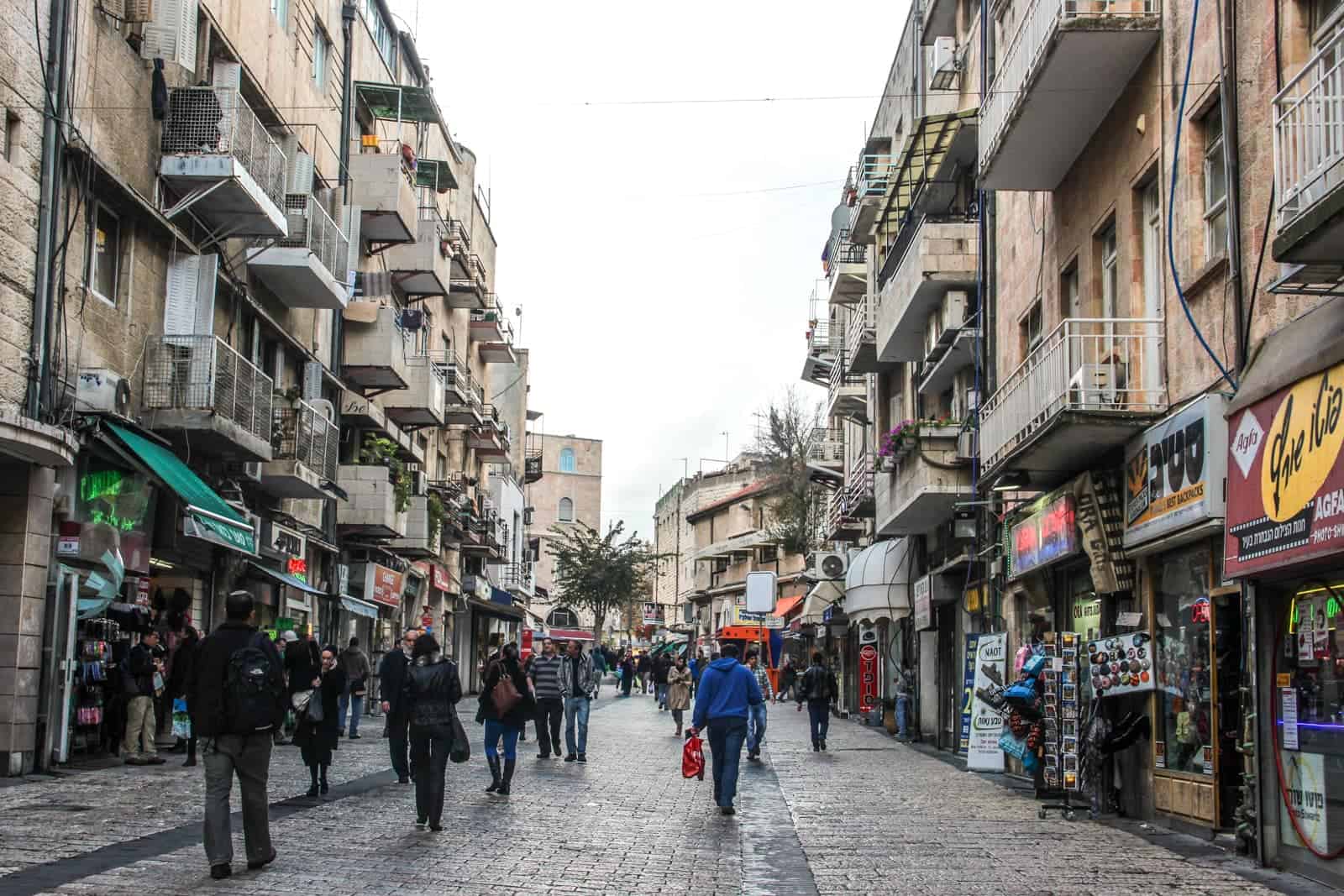 People walking through a busy shopping in Jerusalem modern city. Both sides of the street are lined with shops, and above each one are floors with metal balconies. 