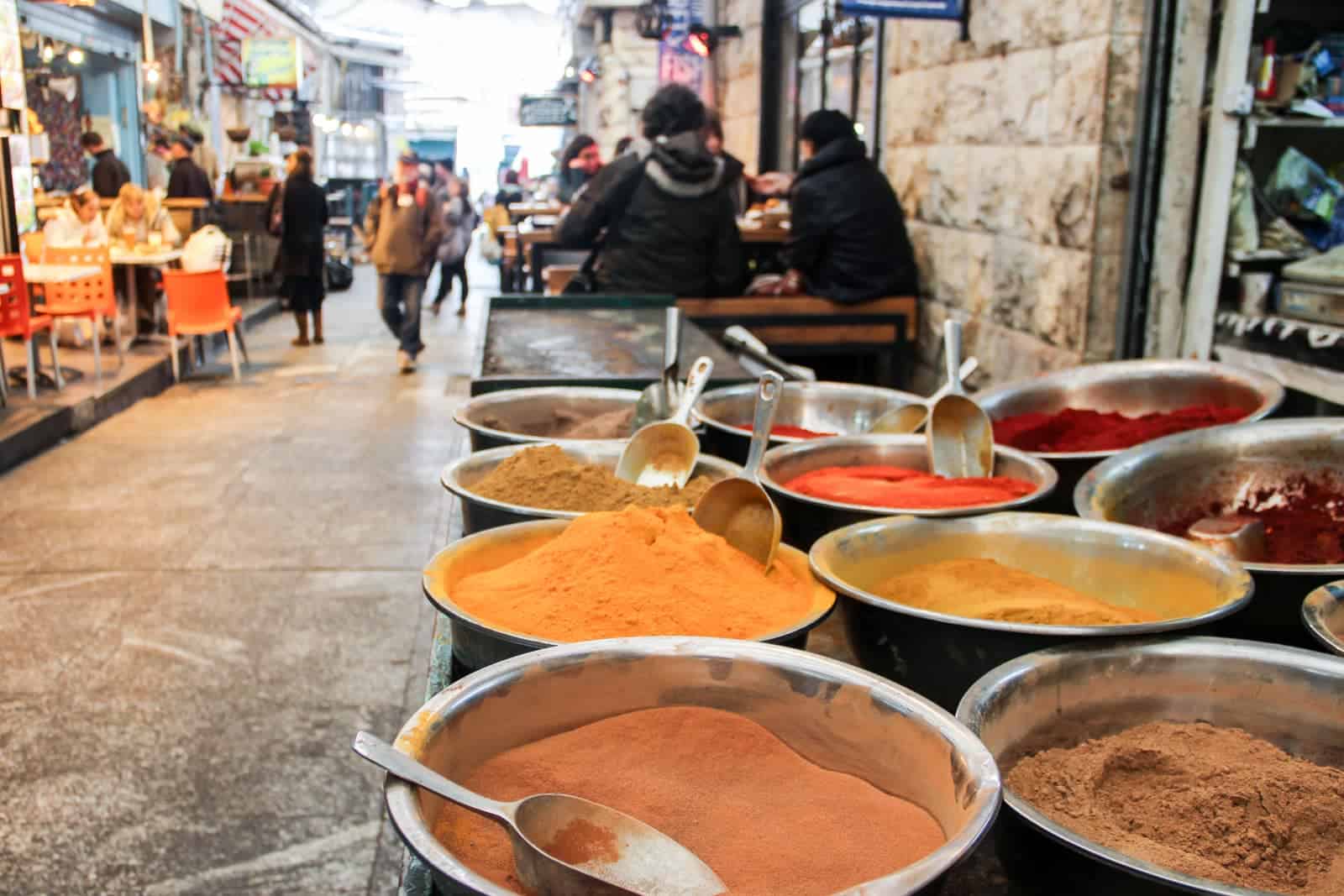 10 metals bowls containing a variety of spices in hues of yellow, orange, red and brown at Mahaneh Yehuda Market in Jerusalem. A group of people are sitting at a table in the background. 