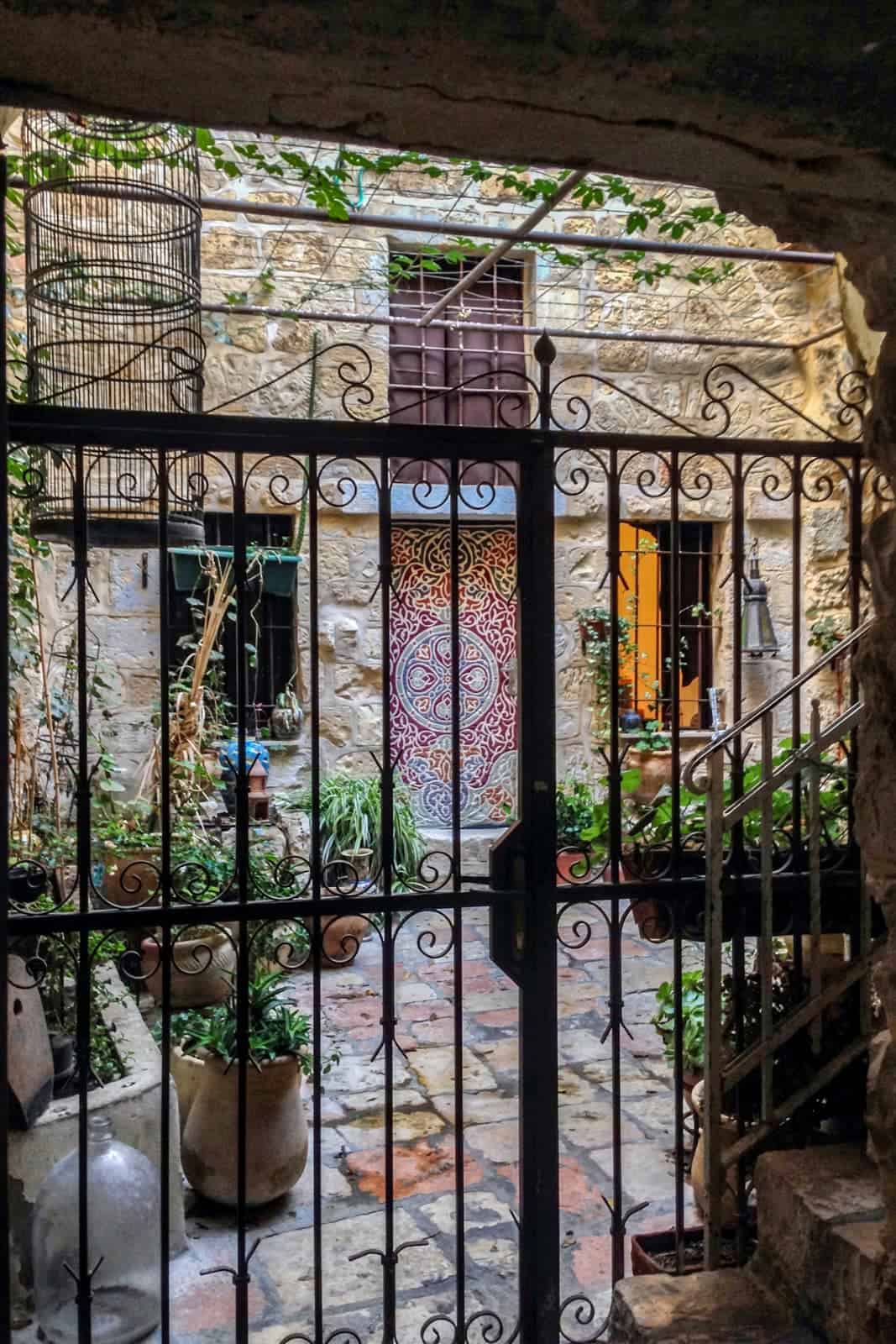 The view through a metal gate to a home in Jerusalem's Old City Armenian Quarter, with a colourful mural door and a courtyard full of potted plants. 