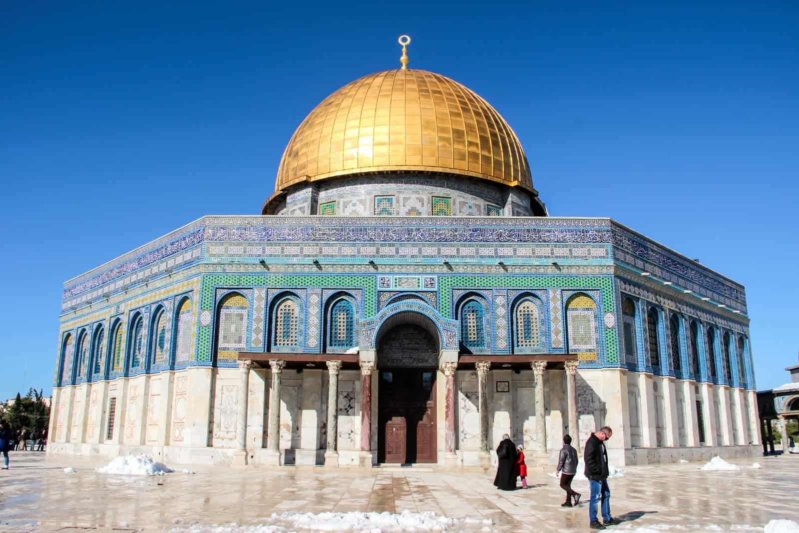 Four people stand outside the columned structure and green, blue mosaic design of Temple Mount in Jerusalem, Israel. The gold dome roof is most recognisable. 
