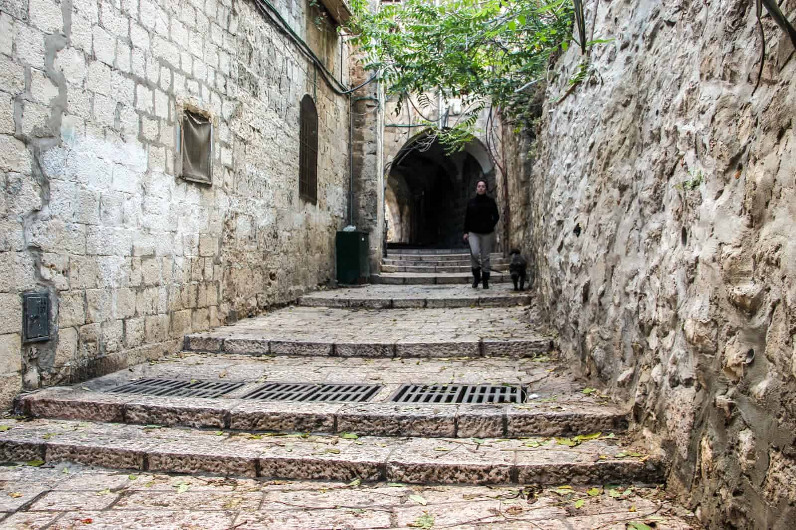 A woman in dark clothing walks down wide stone steps with her dog, in a stone-walled alleyway in Jerusalem Old City in Israel. 
