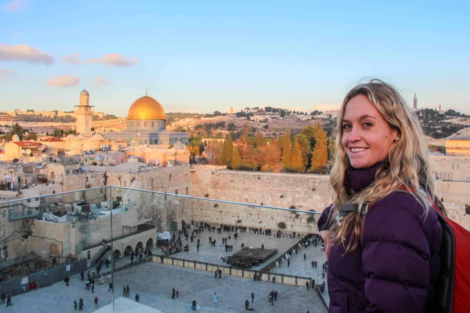 A woman looks at the camera standing at a see-through railing. Behind her and below is the yellow walls of Jerusalem Old Town and people gathering at the tall Western Wall. In the far background a golden dome shines in the sunlight next to a small tower. 