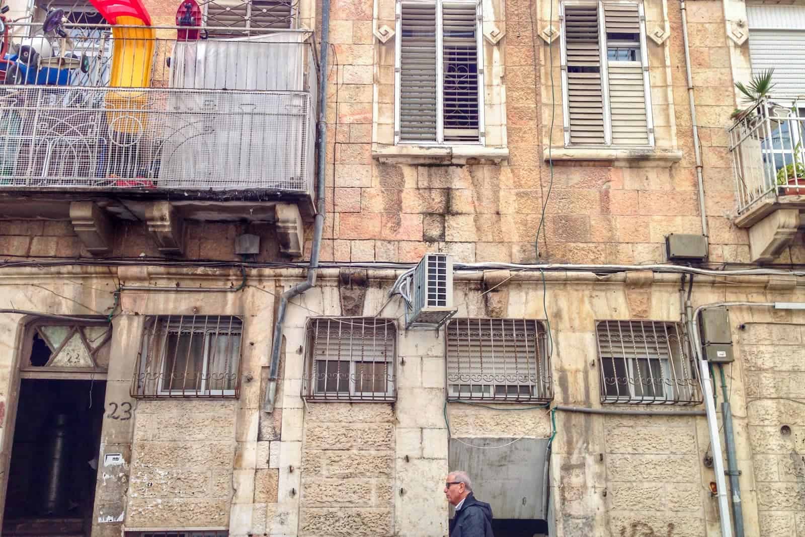 A man walks past a beige brick building in Jerusalem. The windows have metal bars and the first floor has a metal balcony.