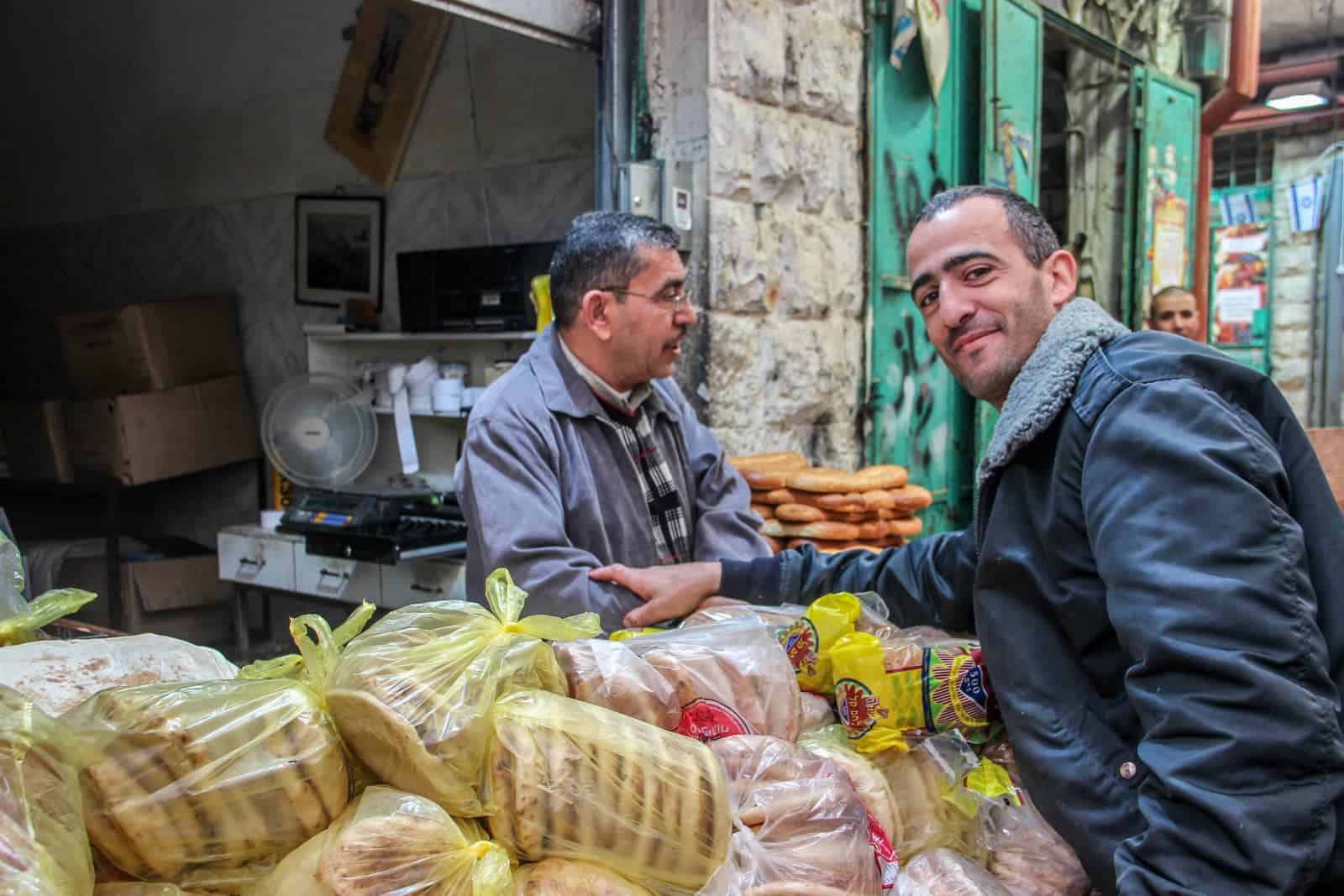 A man at a stand filled with bread in yellow bags, touches the arm of the male vendor affectionately. The door behind them in mint green and part of an old stone wall. 