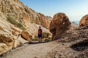 A woman stands at the end of a long dusty path, between a high golden rock face to the left and a large round rock to the right. The golden rock landscape of Ein Gedi Nature Reserve in the Israel desert.