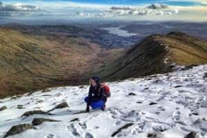 A woman with blue jeans and an ornage backpack, crouches on the snow atop of a mountain in England's Lake District. Behind here is a wide view of the soft, curves of a green valley and beyond that, a lake that seems to be sitting just under the sky.