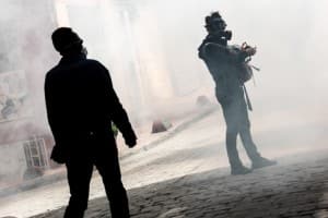 Too men, darkened by the shadows, in rioot gear and gas masks, standing within a cloud of tear gas on the street.