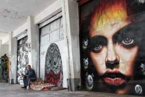 A man sits on a blanket in a white alleyway, filled with blocks of street art. The most prominent mural is one of a woman's face on a black background.