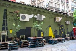 A grass green coloured building with street art depicting black buildings with yellow lights. Woden chairs and tables stand outside the building and the street is empty of people.
