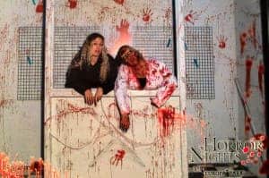 A woman stands in a white box next to an actor dressed as a blood-covered zombie.