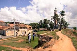 A white lighthouse stands at the end of a coastal peninsula of golden sand and green grass. People can be seen standing next to and walking past the low rise orange building on the left.