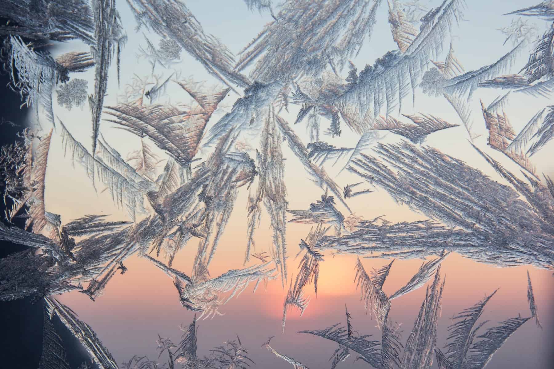 Leaf-like patterns formed by frozen water make a pattern on a window that looks out towards an soft orange sunset. 