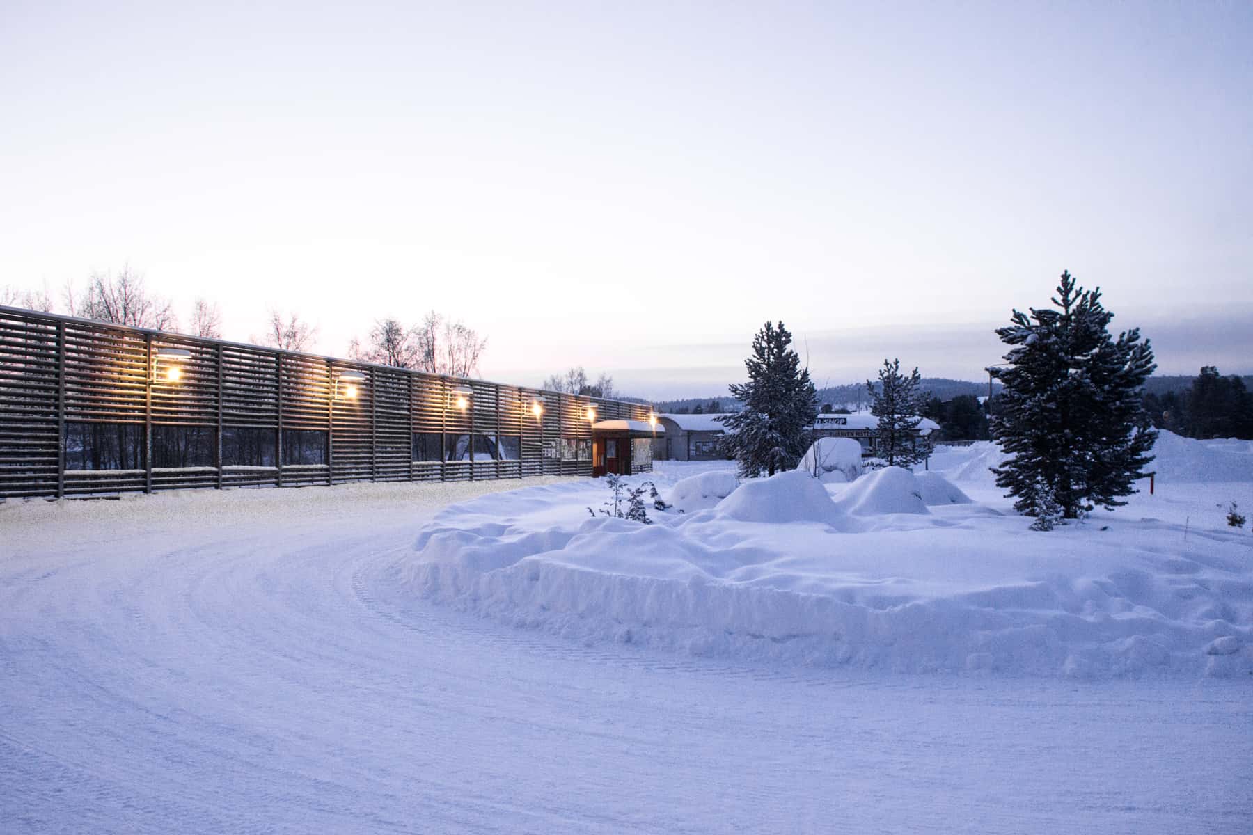 A low, long rectangular wooden building next to a snow covered curved road and a roundabout of snow.