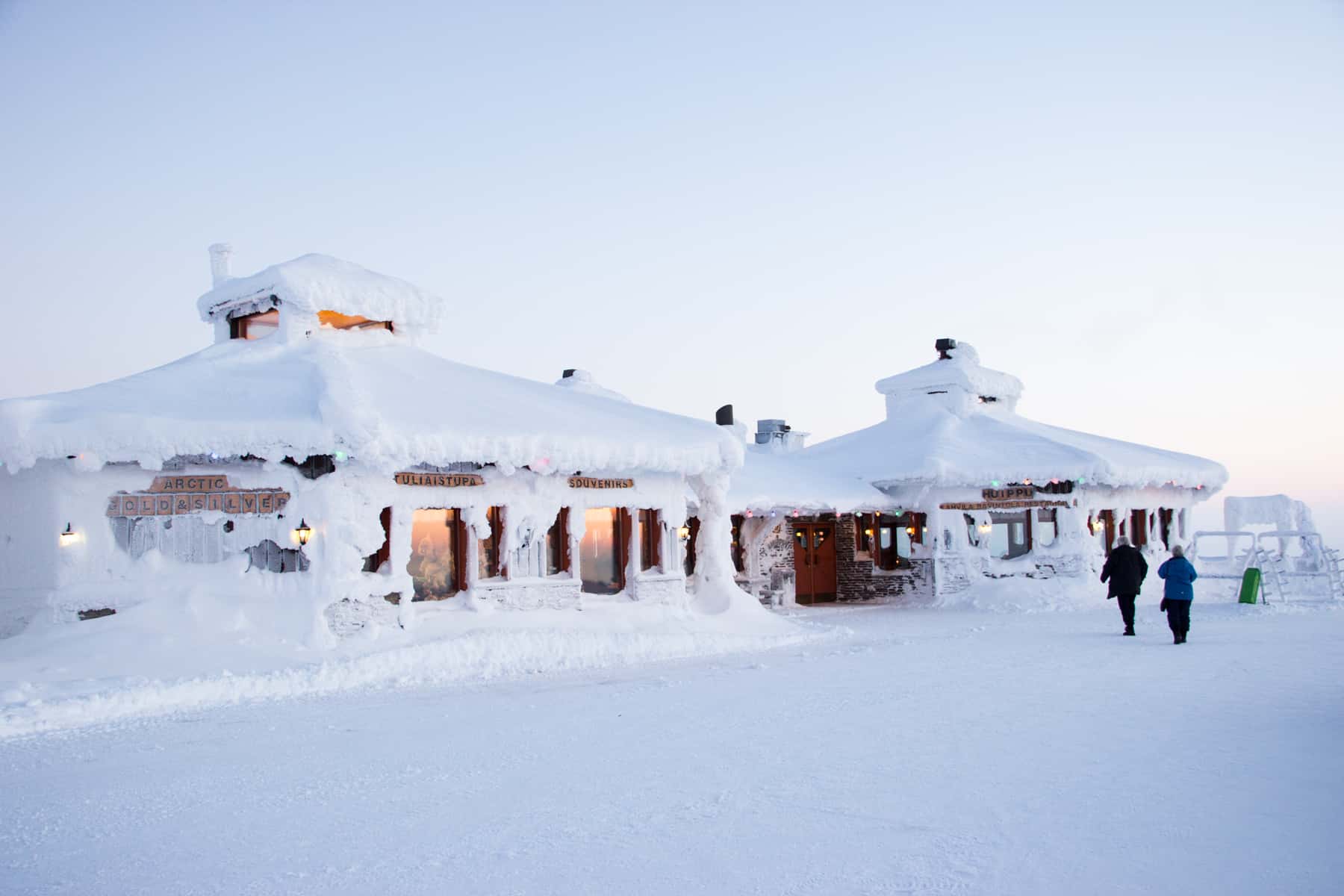 Two people walk towards two octagonal buildings with two-tiered roofs covered in bright white snow under a bright white sky. Only a few windows are visible. 