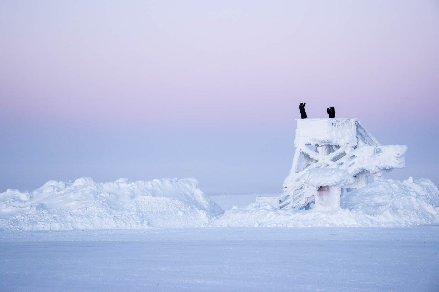 Two people dressed in black stand upon a viewing platform covered in snow and ice in a flat icy wilderness under a pink and purple sky.