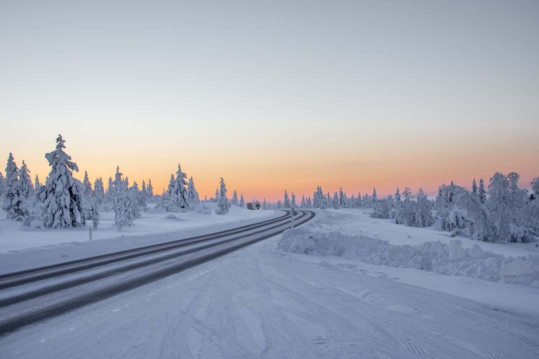 A narrow gravel road cuts through a snow covered forest and curves to the left towards an orange sunset that only lights up half the sky. 