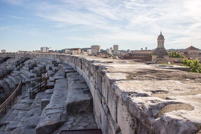 Picture of the top wall of a white stone Roman Ampitheatre, with part of the skyline of Nimes city in France in the background.