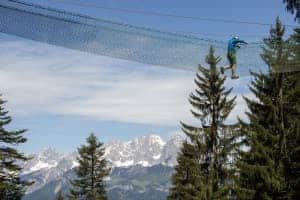 A man walks through a net canope between trees to a backdrop of mountains, at a climbing park in Tirol, Austria.