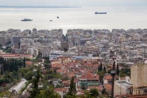 An elevated view looking down on the city of Thessaloniki in Northern Greece. The landscape changes from clay coloured buildings in the foreground, to modern silvery white blocks in front of the sea, which has ships passing.