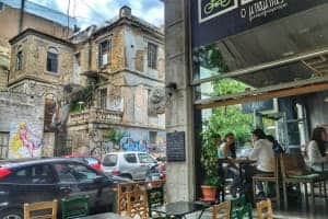 People sitting in a cafe on a street lined with cars and buildings covered in graffiti.