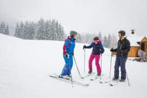 A ski instructor (in blue) talking to two people on a small snow-covered hill in front of a wooden hut.