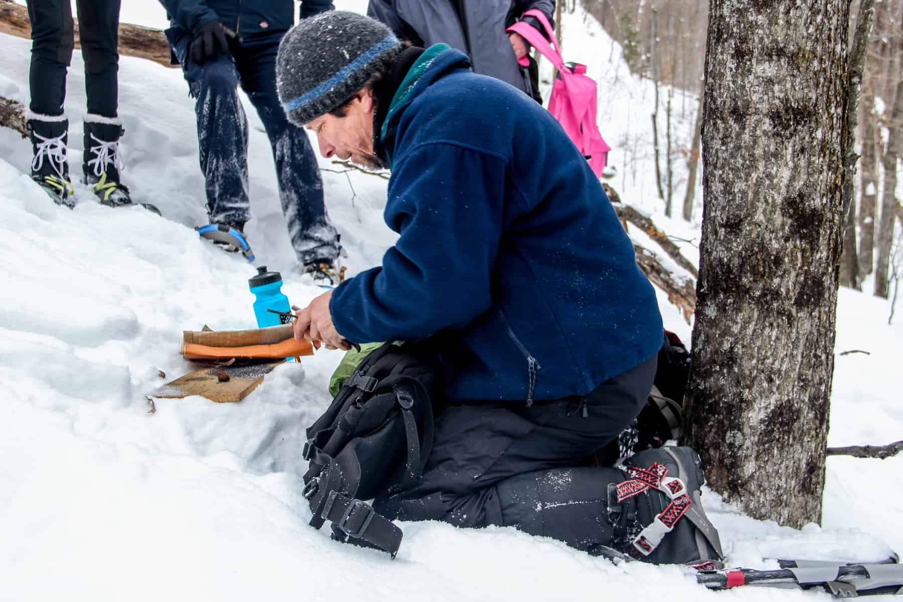 A man, on the knees, works with bark and paper trying to light a fire in a snow covered forest.