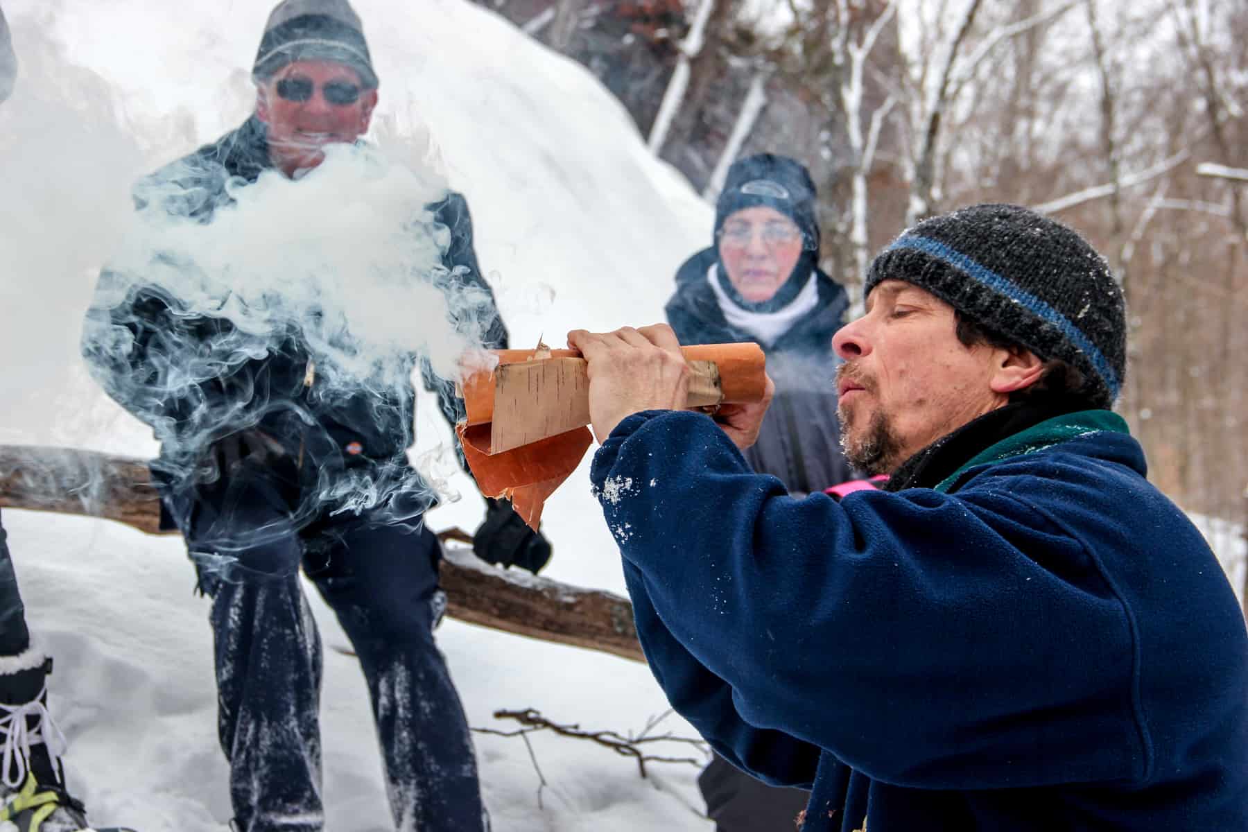 Watched by two people, a man blows into paper releasing a bellow of smoke in a snow drenched forest.