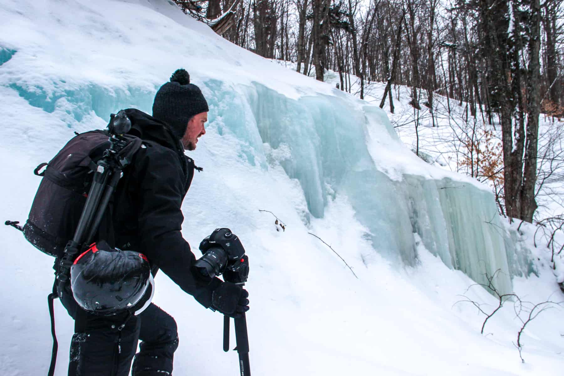 A man in black clothing, carrying camera gear and a helmet, walked past a blue-ice glacier in a snow covered forest in Mont Tremblant in winter. 