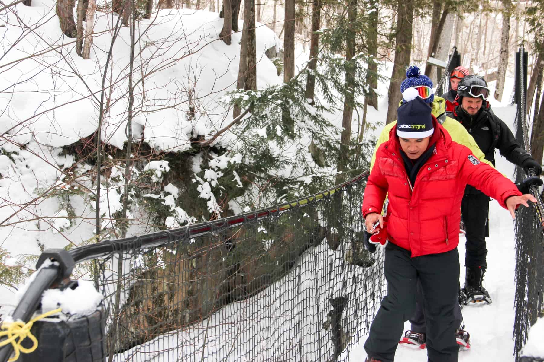 Four people wearing bright coloured ski gear snowshoe in Mont Tremblant, crossing a metal bridge in a forest. 