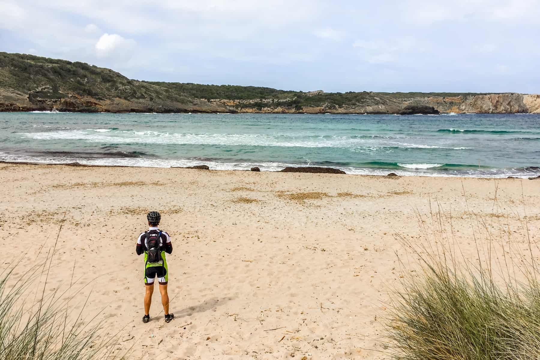 A man in cycling clothing stands on a white sand beach looks out to the green-blue sea backed by low green hills. 