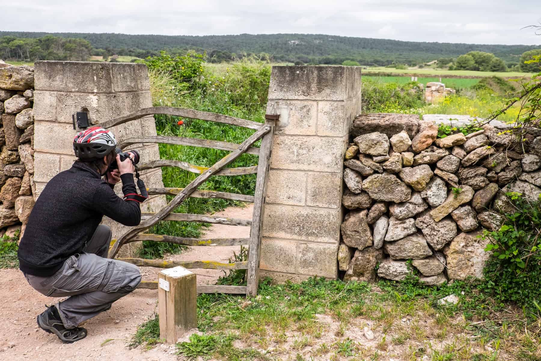 A man kneels and takes a picture of a green field through the openings of a wooden gate. 